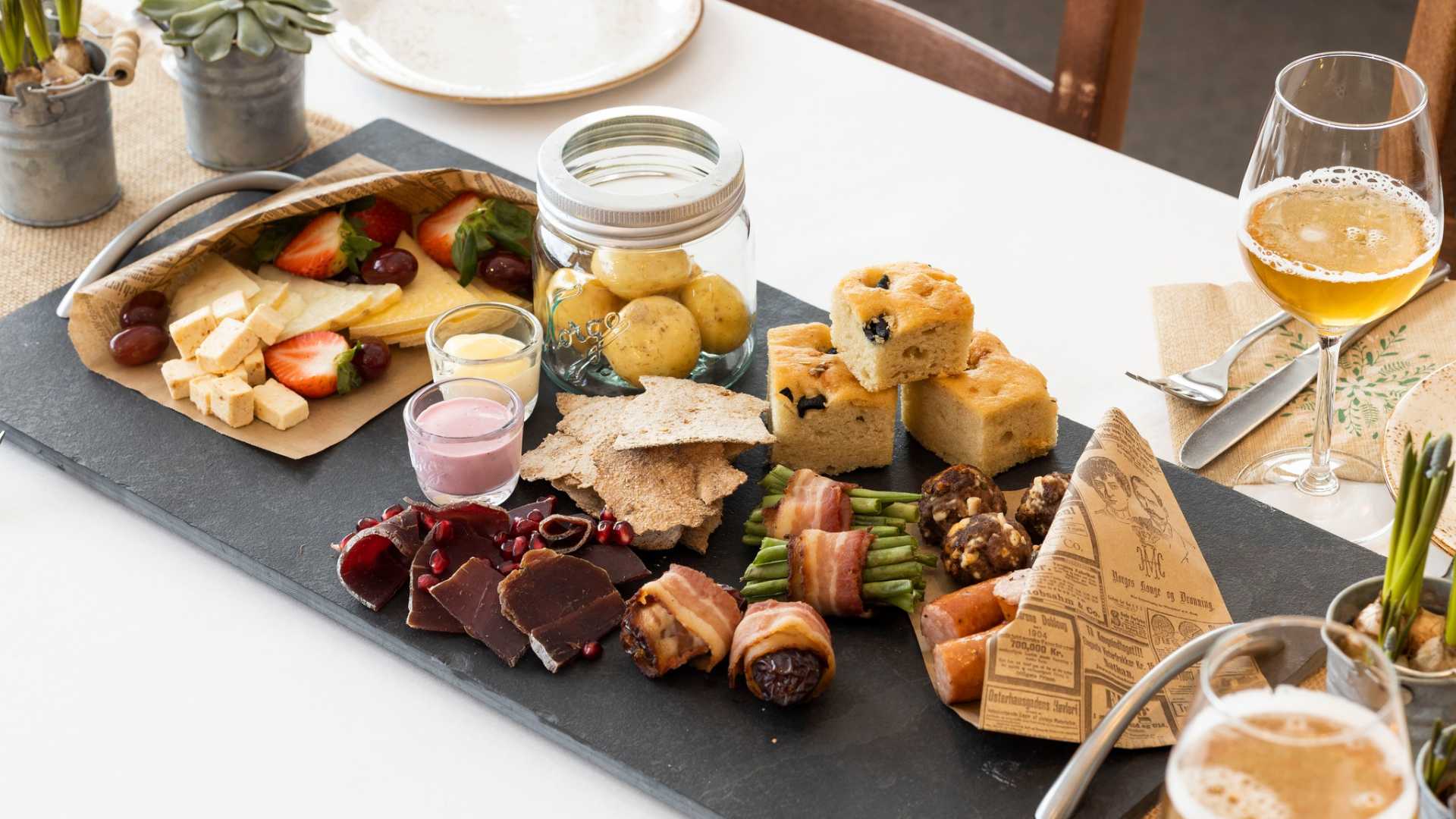 Assortert delikatessefat med ost, frukt, baconruller, brød og dip på skiferplate, servert med øl.