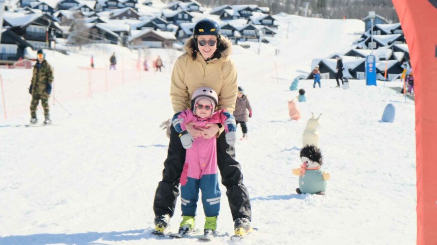 Voksen og barn på ski i solfylt skianlegg, omringet av snødekte hytter og leker. Vinterglede i fjellet.