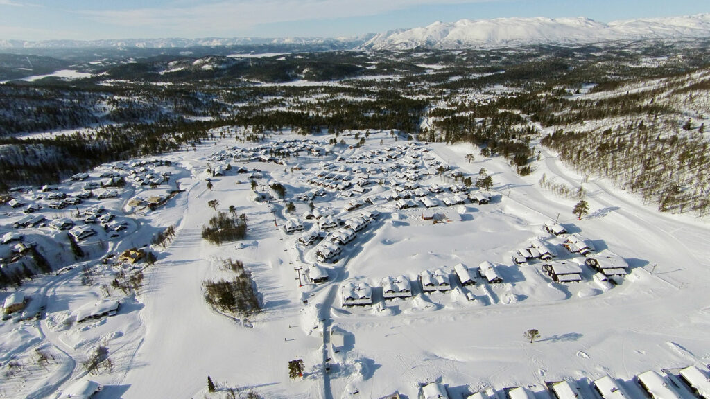 Snødekt landsby i fjellene, med hytter og skog, sett fra luften på en solfylt vinterdag i Norge.