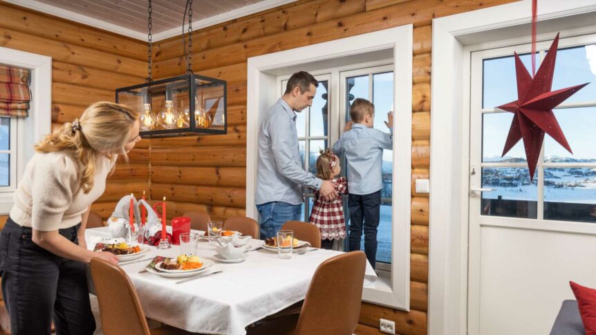 Familie ser ut av vinduet i julepyntet hytte, mens mor dekker bordet med julemiddag.