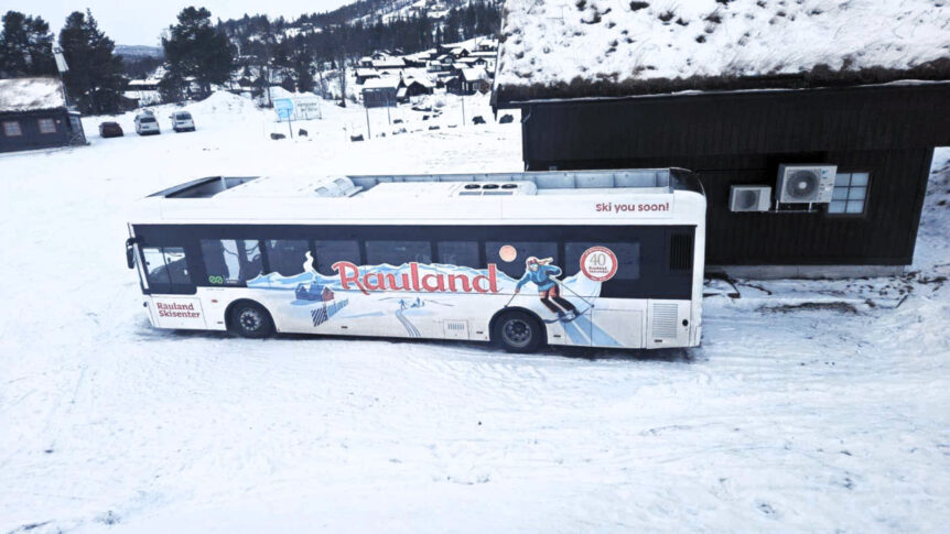 Buss med Rauland Skisenter-reklame parkert i snødekt landskap nær hytte.
