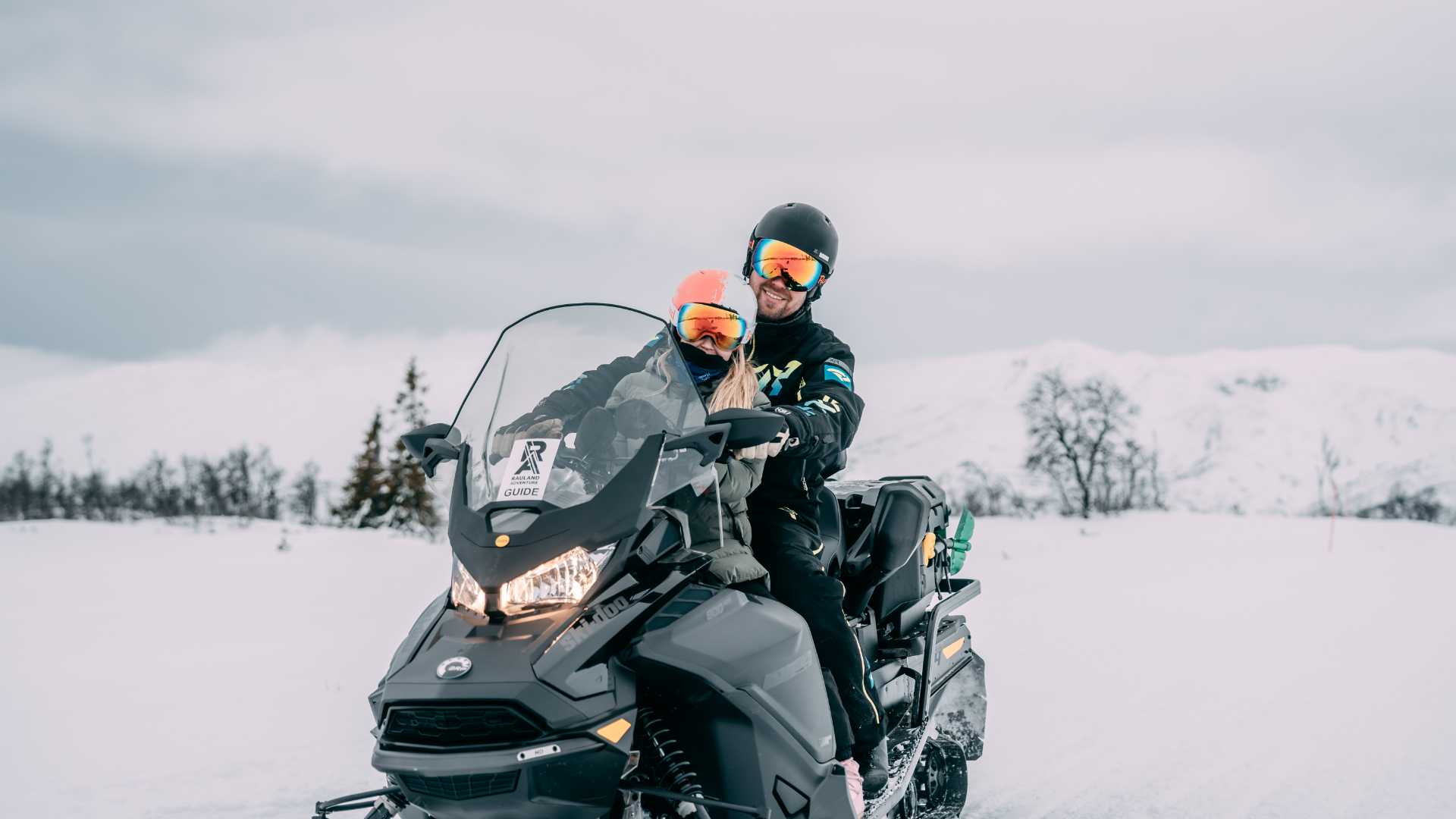To personer kjører snøscooter i snødekte fjelllandskaper, iført fargerike skibriller og hjelmer.
