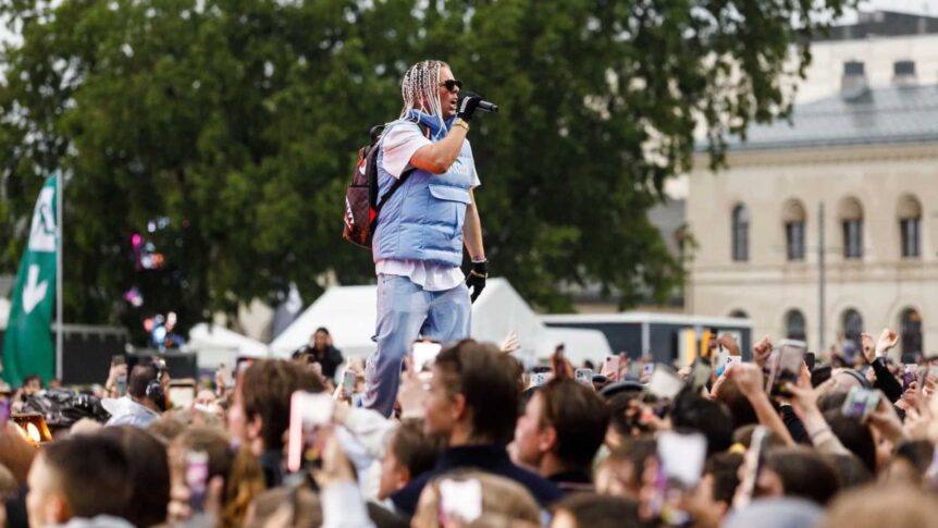 Kunstner i blå vest synger på scenen foran et stort publikum under en utendørskonsert i en park.