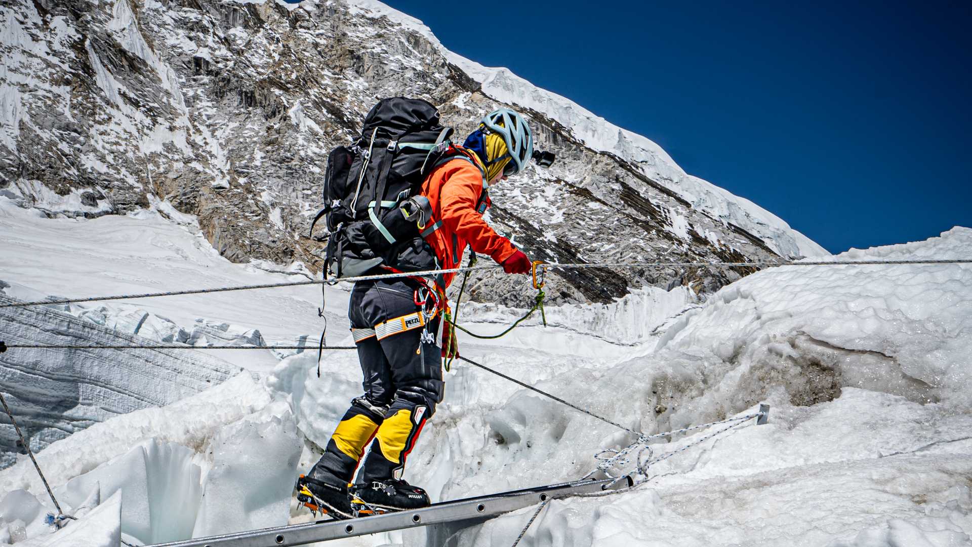 Fjellklatrer krysser isbro med ryggsekk og hjelm, omgitt av snødekte fjell og blå himmel.