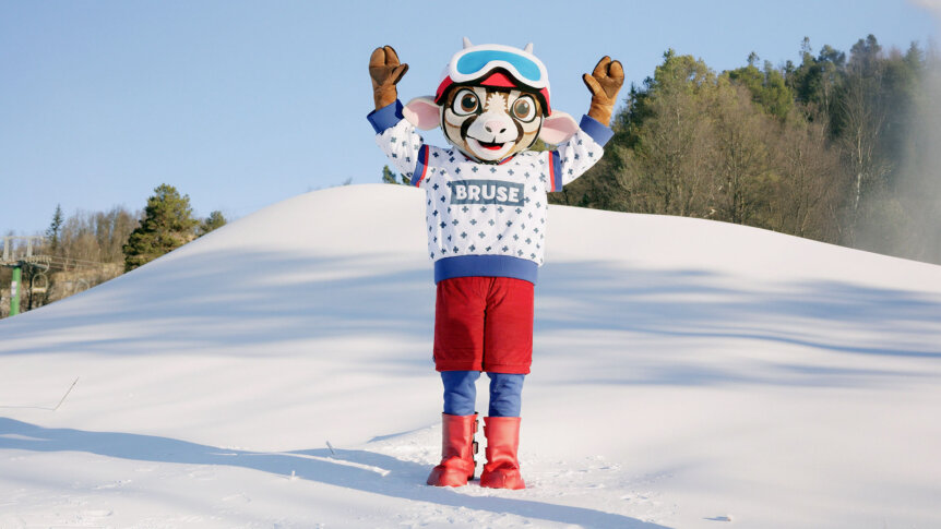 Maskot i vinterklær jubler på snødekt område med skog i bakgrunnen.