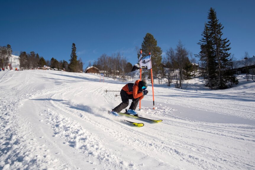 Person som står på ski i høy fart ned en snødekt bakke under klar blå himmel.