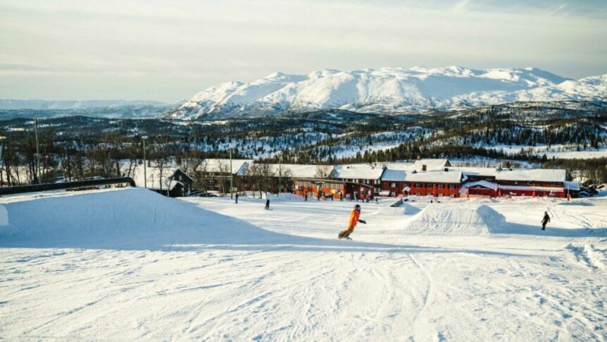 Snowboarder i et norsk skianlegg med snødekte fjell og trebygninger i bakgrunnen. Klar vinterhimmel.