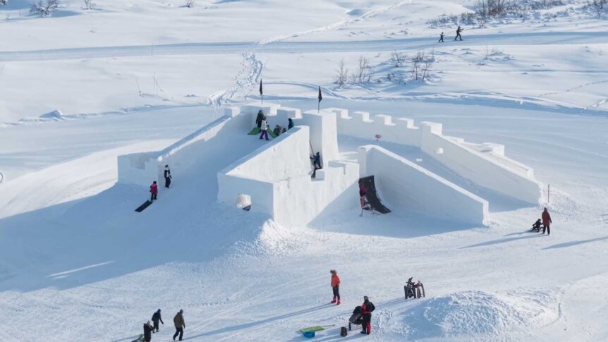 Snøborg med sklier og barn i lek på vinterdag i fjellandskap. Skispor i bakgrunnen.