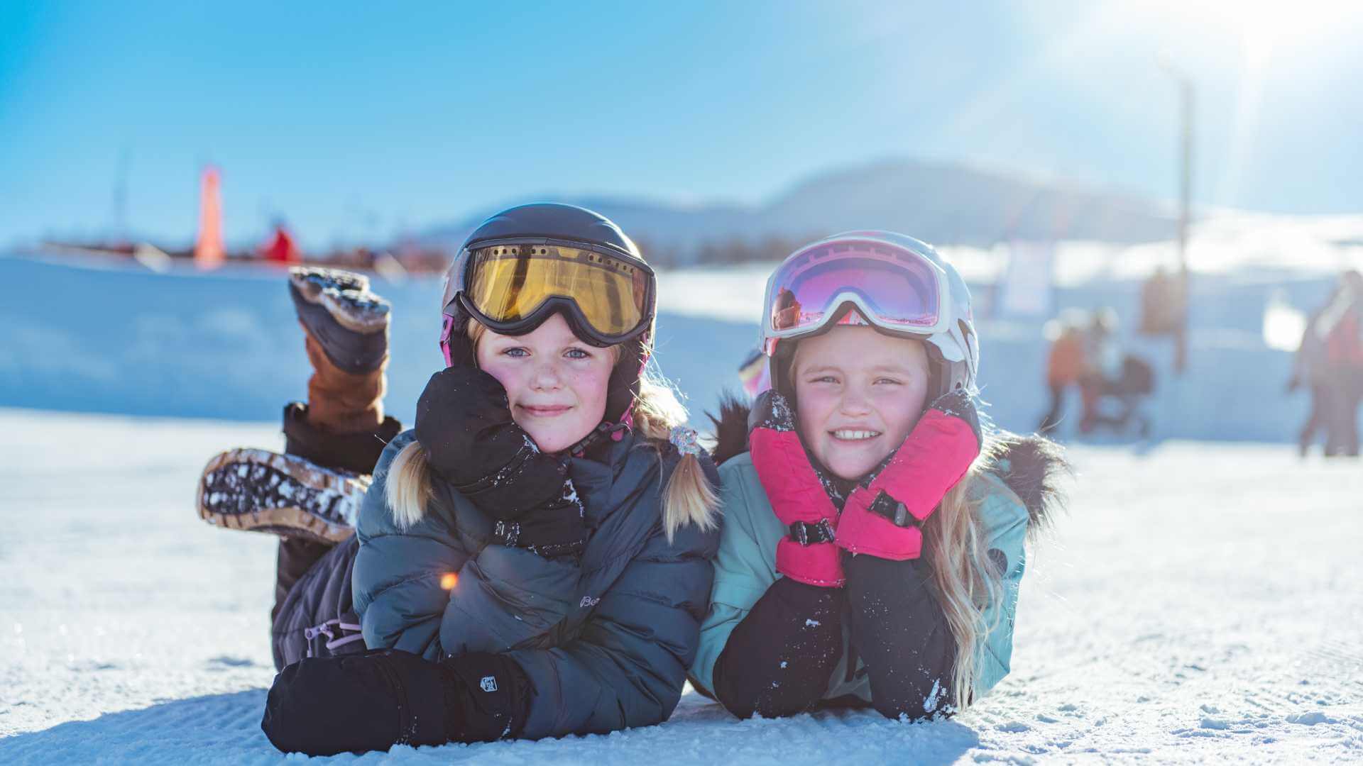 To jenter i skiutstyr ligger på snøen, smilende og klare for vintermoro i solfylt fjellandskap.