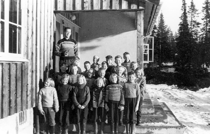 Svart-hvitt bilde av en skoleklasse og lærer står på trappen til en trebygning, omgitt av snødekt landskap og skog.