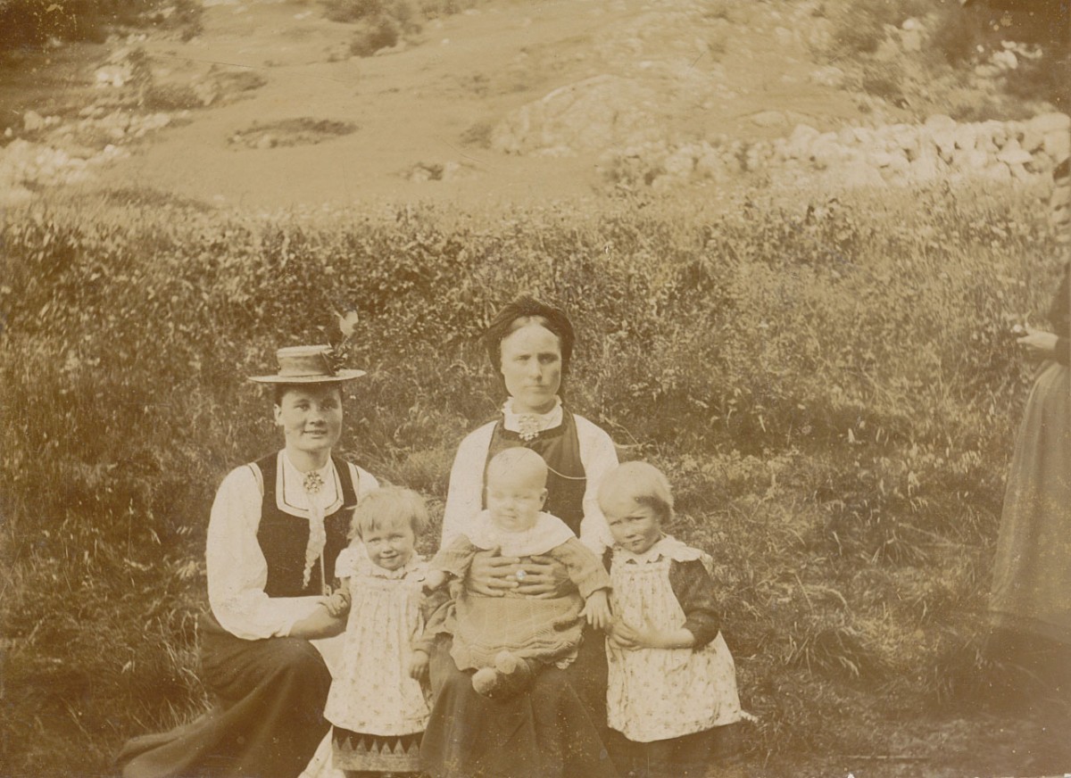 En familie på fem i gammeldags klær poserer i en naturlig setting med gress i bakgrunnen.