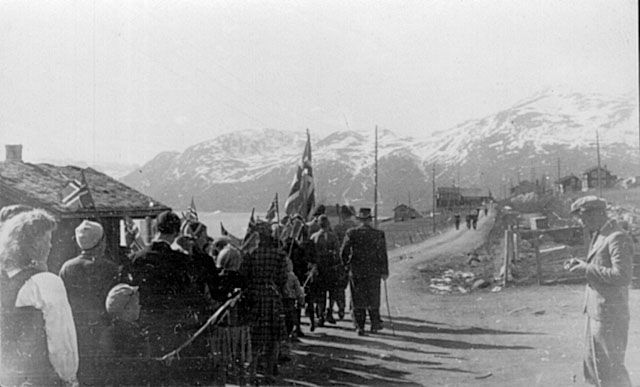 Svart-hvitt foto av marsjerende personer med flagg i norsk bygd, fjell i bakgrunnen, tidlig 1900-tall.