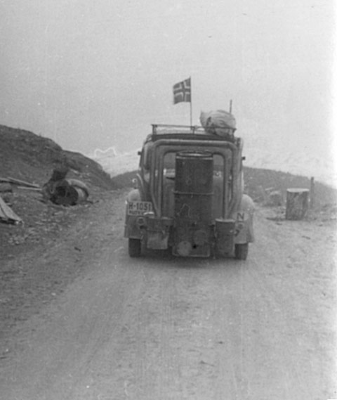 Gammel bil med norsk flagg på landevei, bærer bagasje på taket. Historisk reisebilde fra Norge.