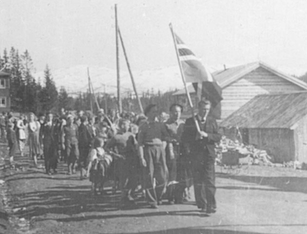 Sort-hvitt bilde av en norsk parade med mennesker som marsjerer og holder et flagg, fjell i bakgrunnen.