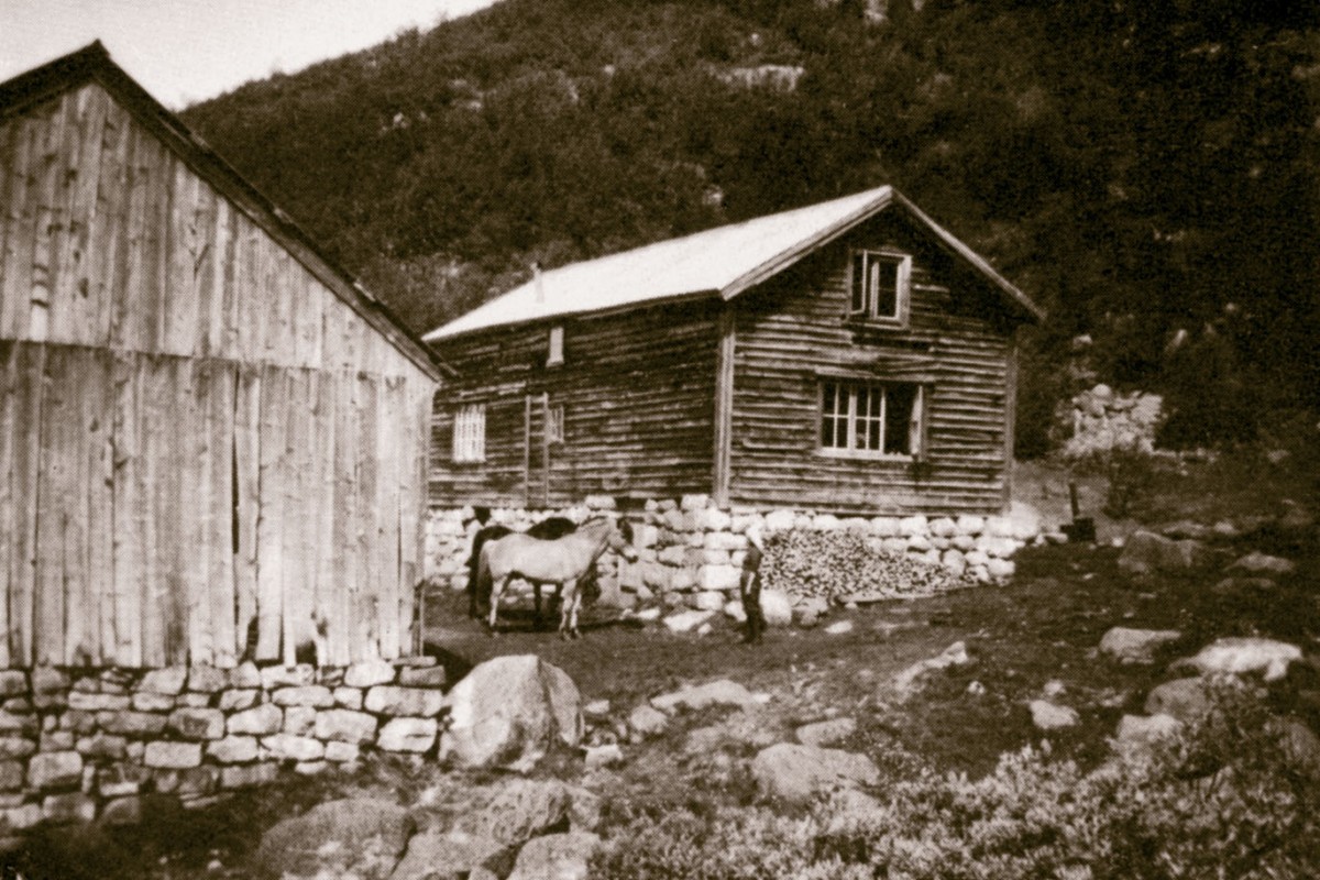 Gammel trebygning med steinmur og hest foran, omgitt av fjellandskap.