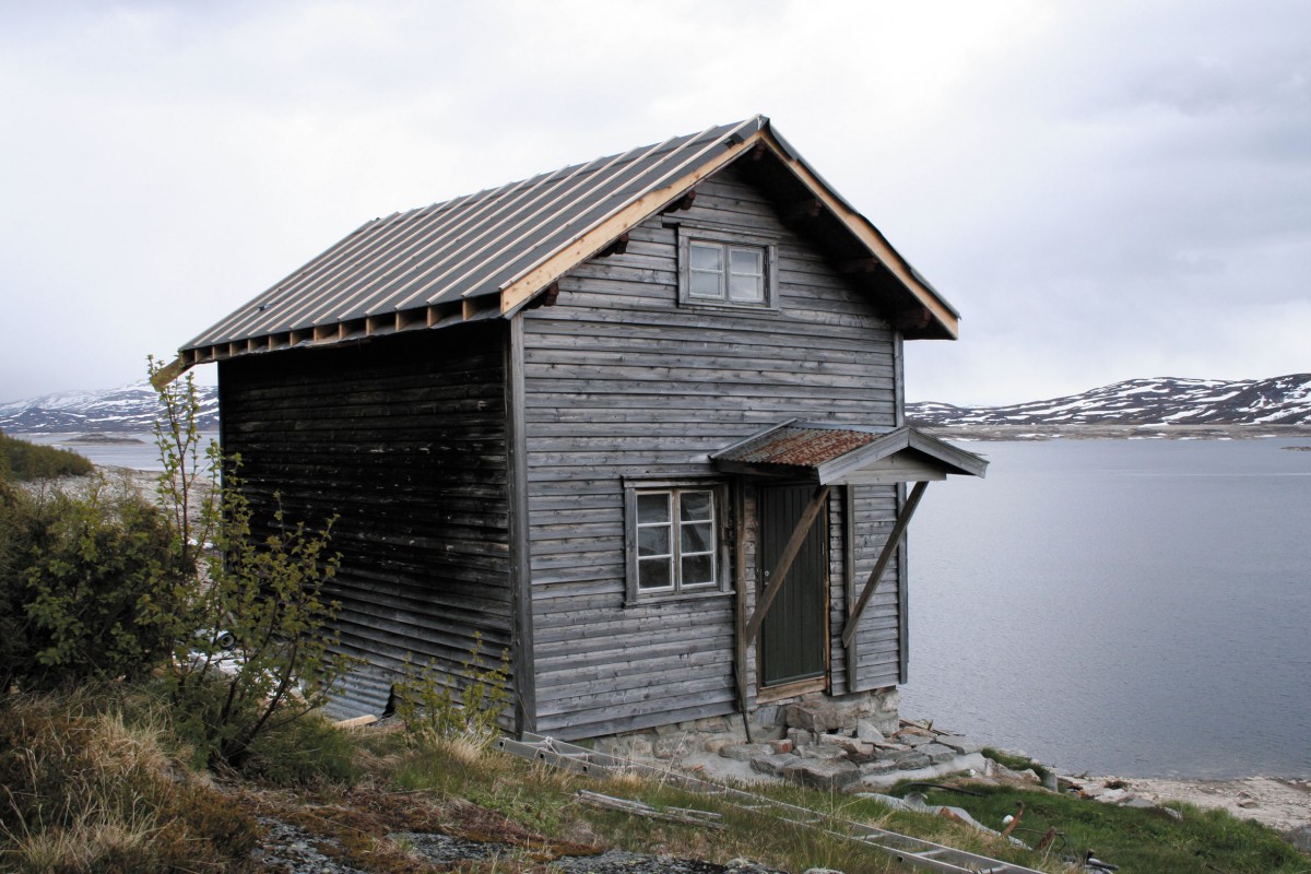 Gammelt trehus ved innsjøen i norsk landskap med fjell i bakgrunnen, under overskyet himmel.