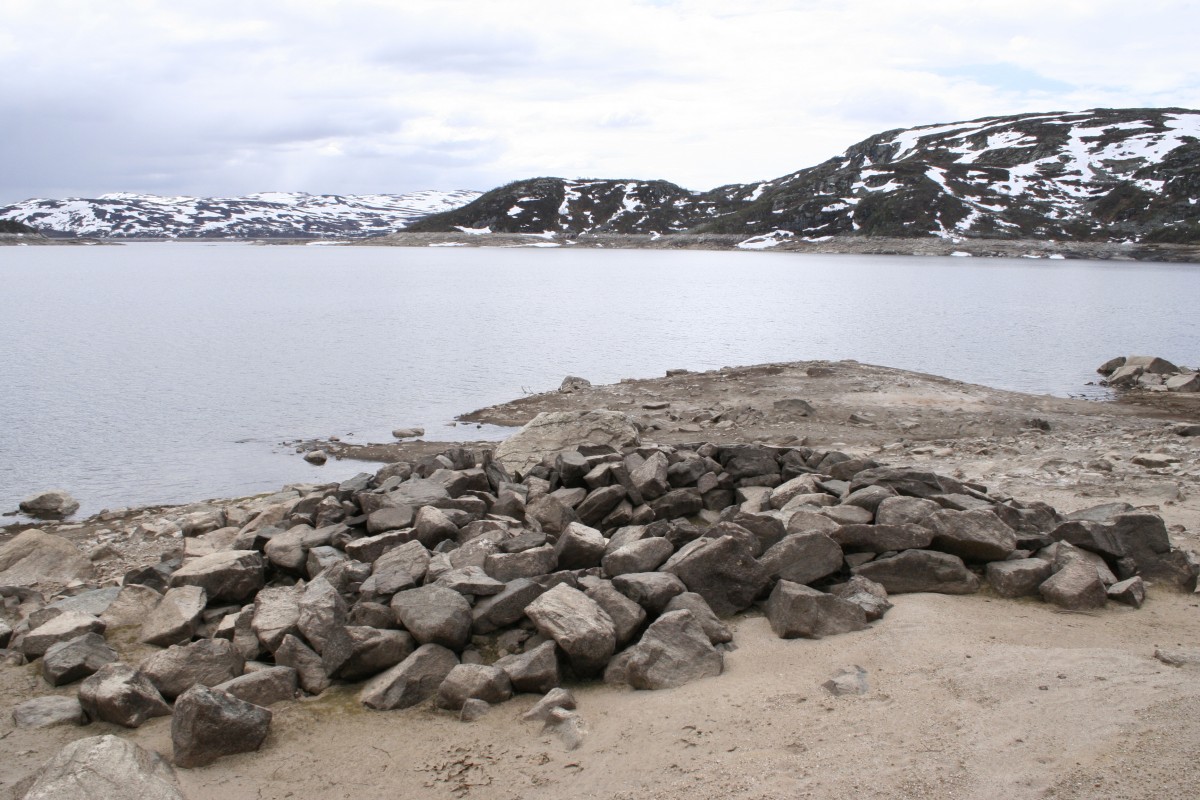 Steinete strand ved innsjø med snødekte fjell i bakgrunnen under overskyet himmel.