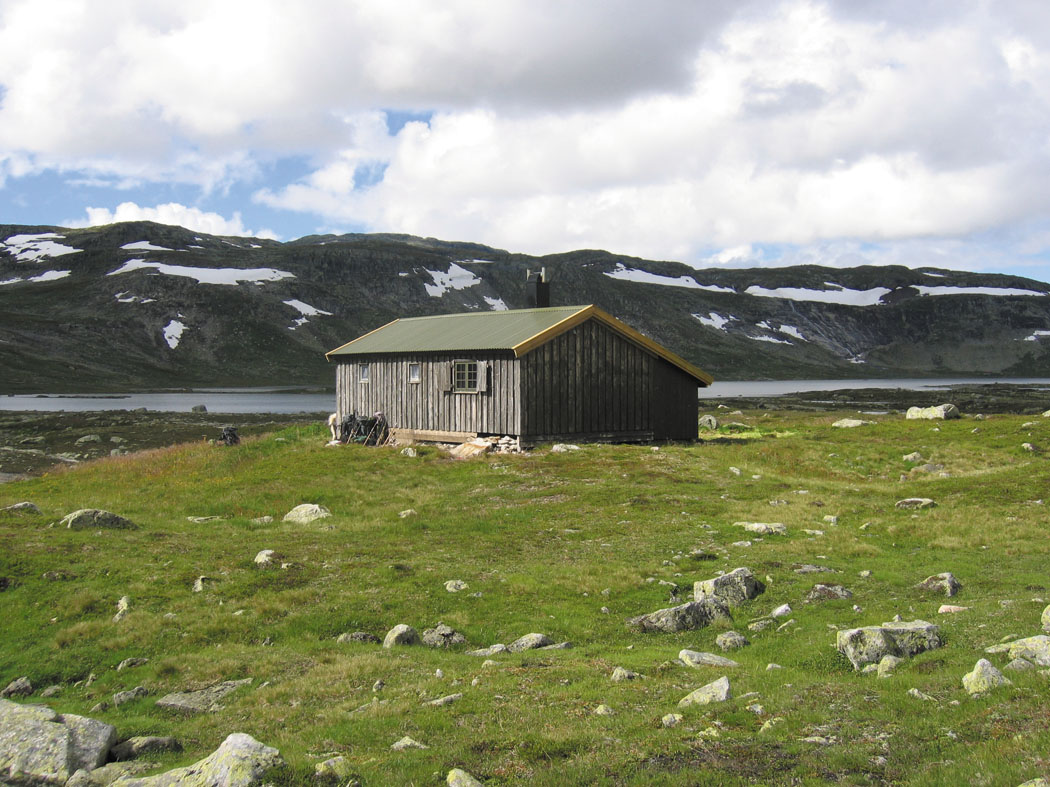 Fjellhytte ved en innsjø med snødekte fjell i bakgrunnen under en skyet himmel.