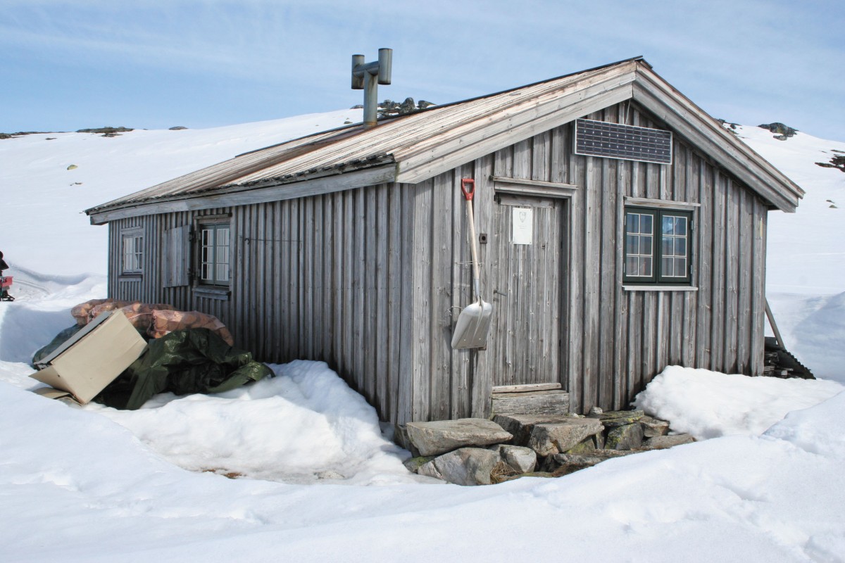 Tre-hytte i snødekte fjell, med solcellepanel på taket og spade hengende ved inngangen.