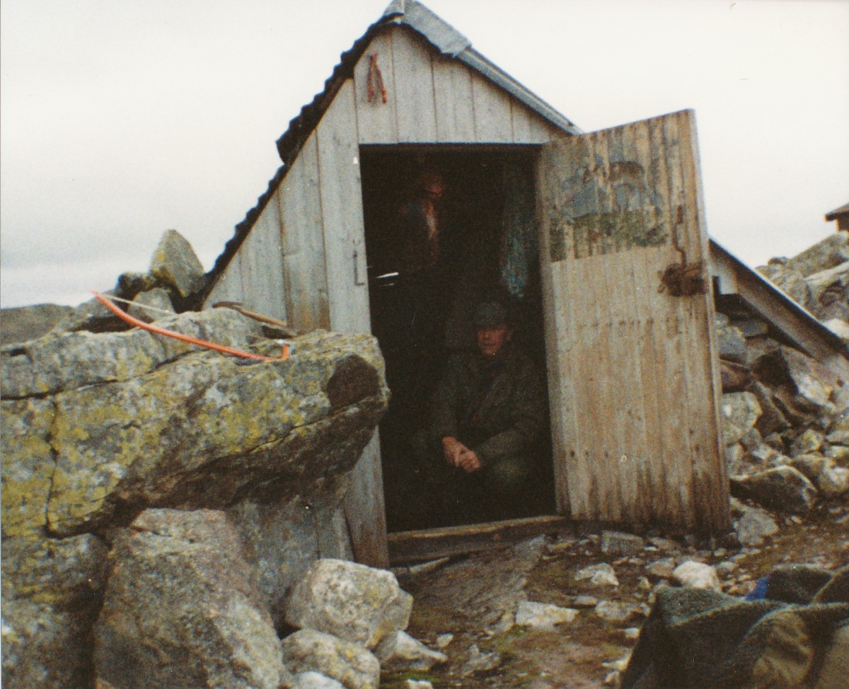 Lite fjellskjul i steinlandskap, mann sitter ved åpen dør. Rustikk atmosfære på norsk fjell.