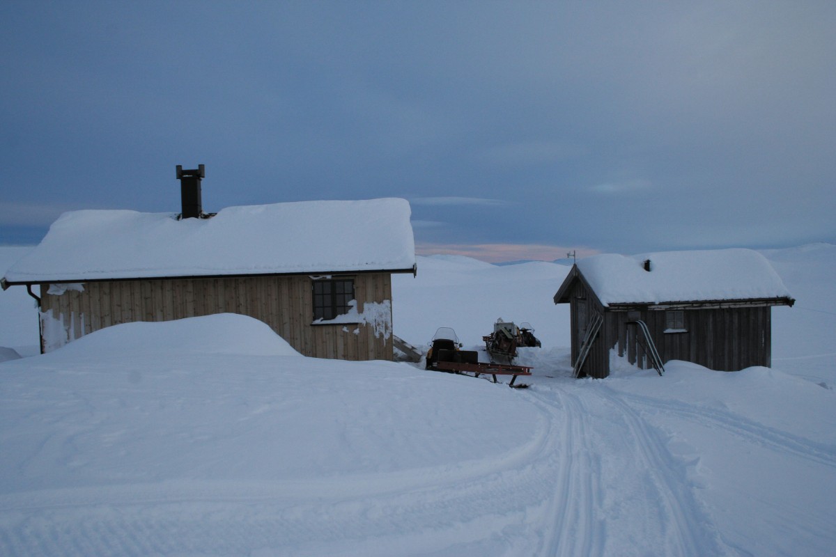 Snødekt fjellhytte og skur med snøscooter ved solnedgang i norsk vinterlandskap.