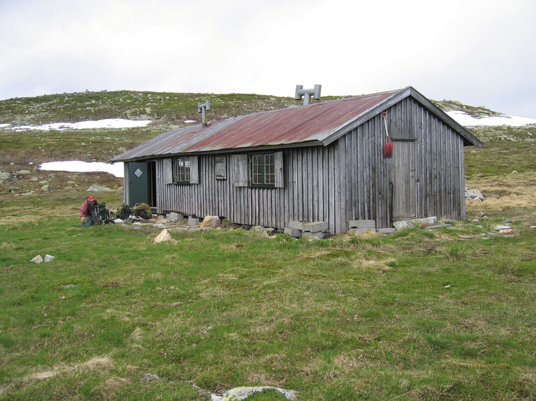 Gammelt trehus i norsk fjellandskap, med gress og snøflekker rundt.