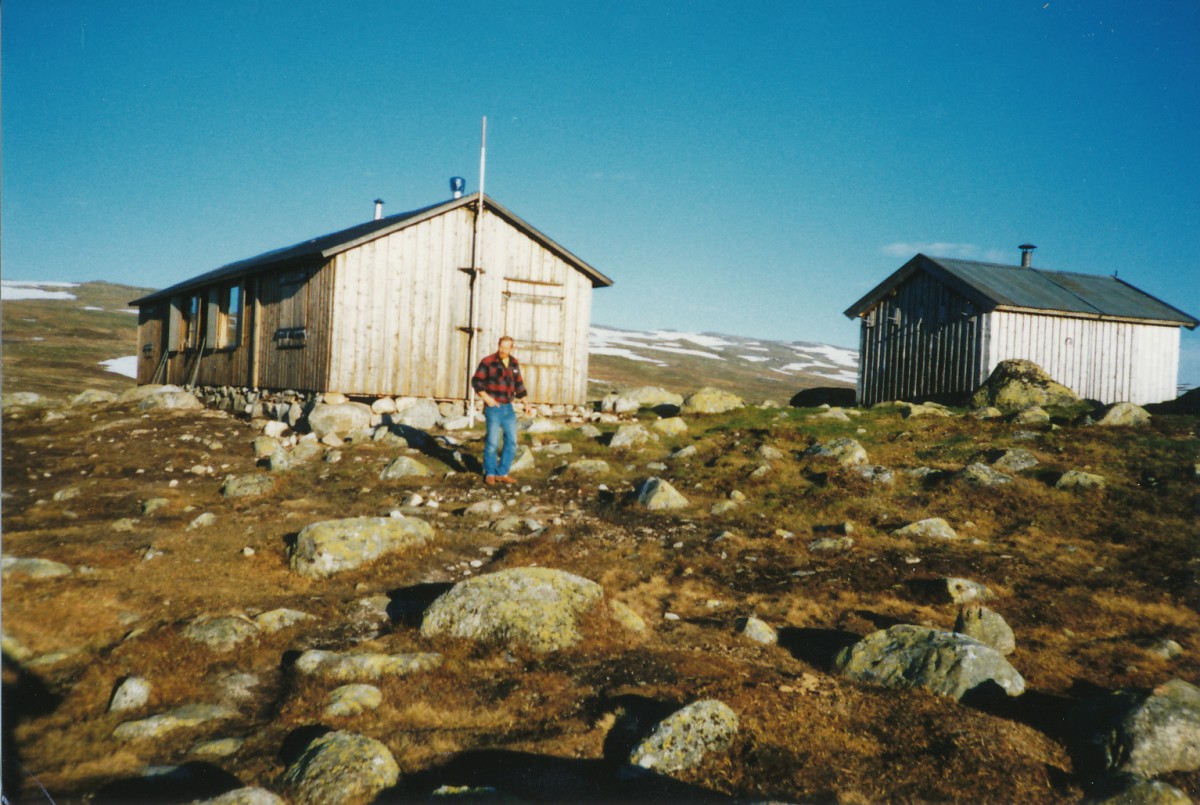 To trehytter i steinete fjellandskap under en klar blå himmel, med en mann i rutete skjorte foran.