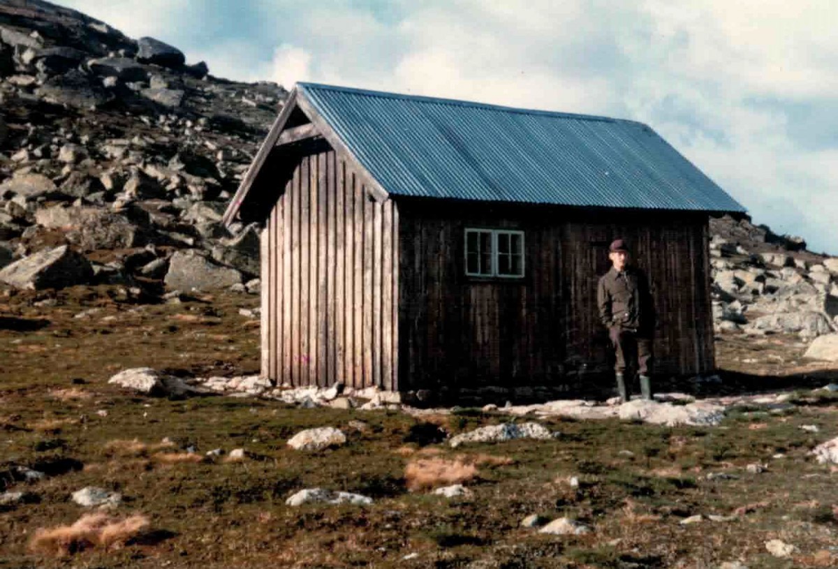 Lite trehus i fjellterreng med person stående ved siden, blått tak og steinete bakgrunn.