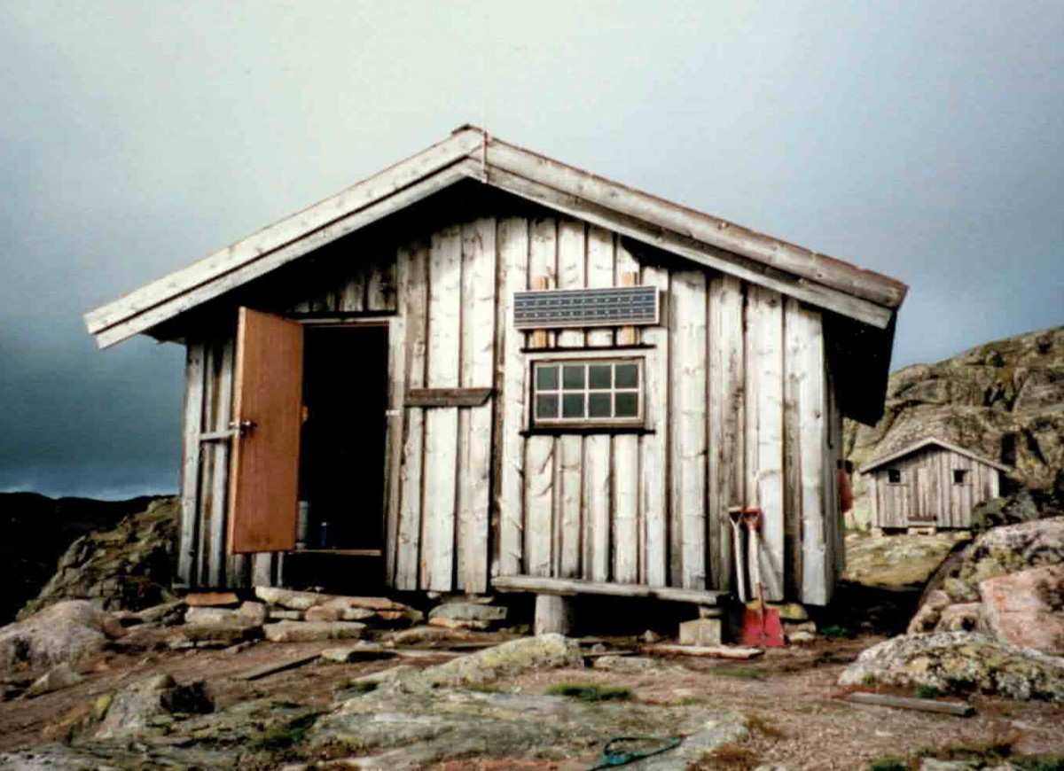 Liten trehytte i fjellet med åpen dør og solcellepaneler, omgitt av steiner og skyet himmel.