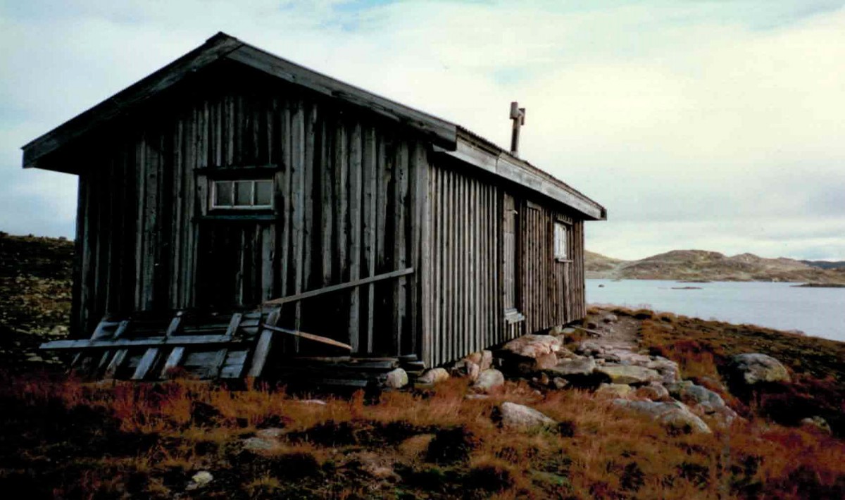 Gammel trehytte ved innsjø i norsk fjellandskap, omgitt av steiner og lyng.