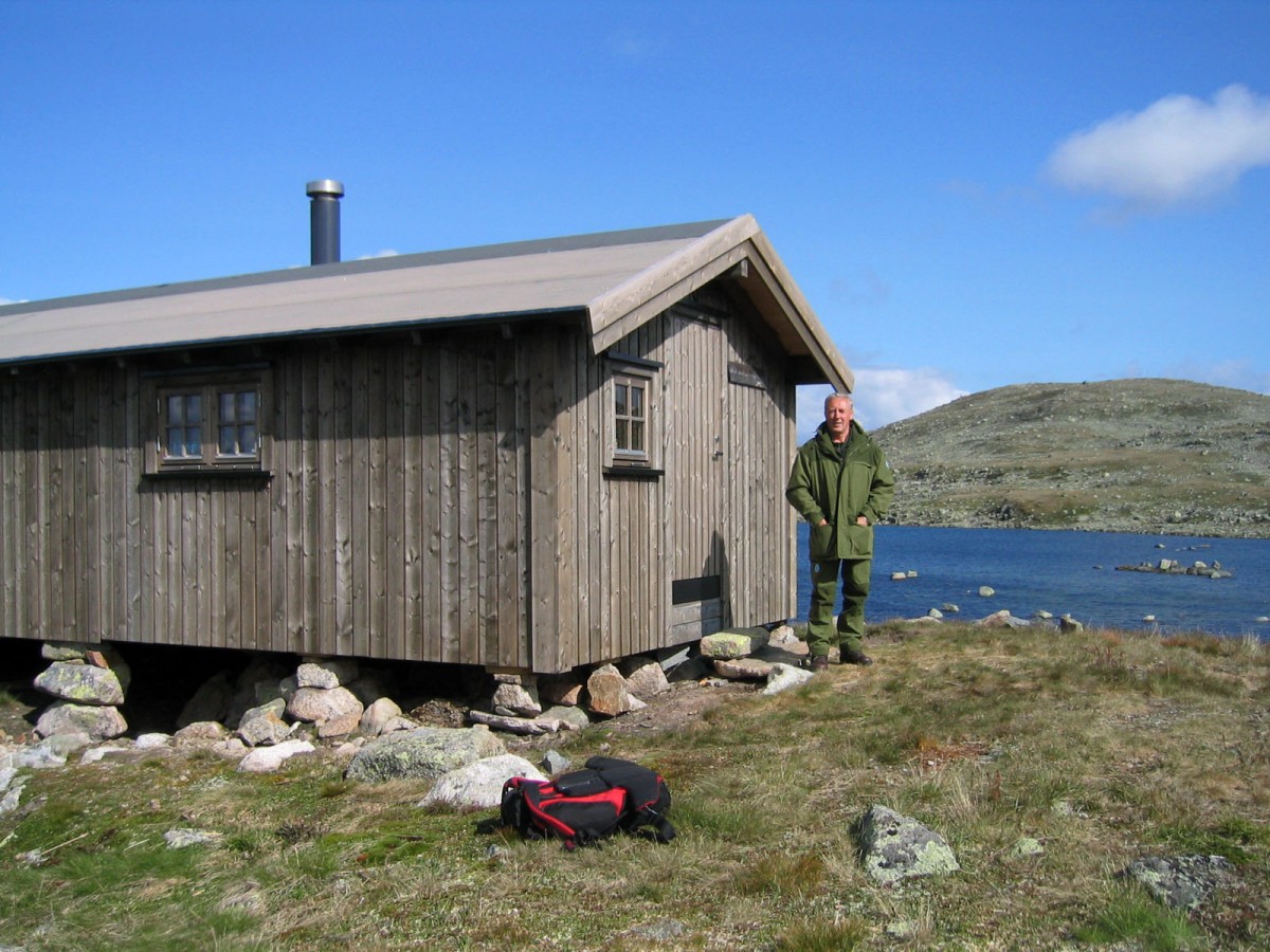 Mann ved fjellhytte ved innsjø i norsk natur, klar blå himmel.