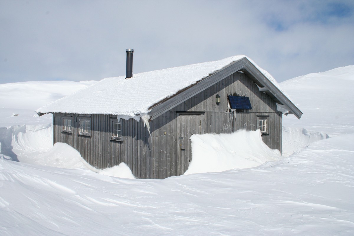 Trehytte i vinterlandskap, dekket av snø i fjellet, solcellepanel på taket.