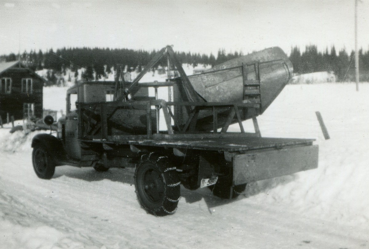 Gammel lastebil med snøkjeder i vinterlandskap, lastet med stor metallbeholder på flatbed, trehytte i bakgrunnen.