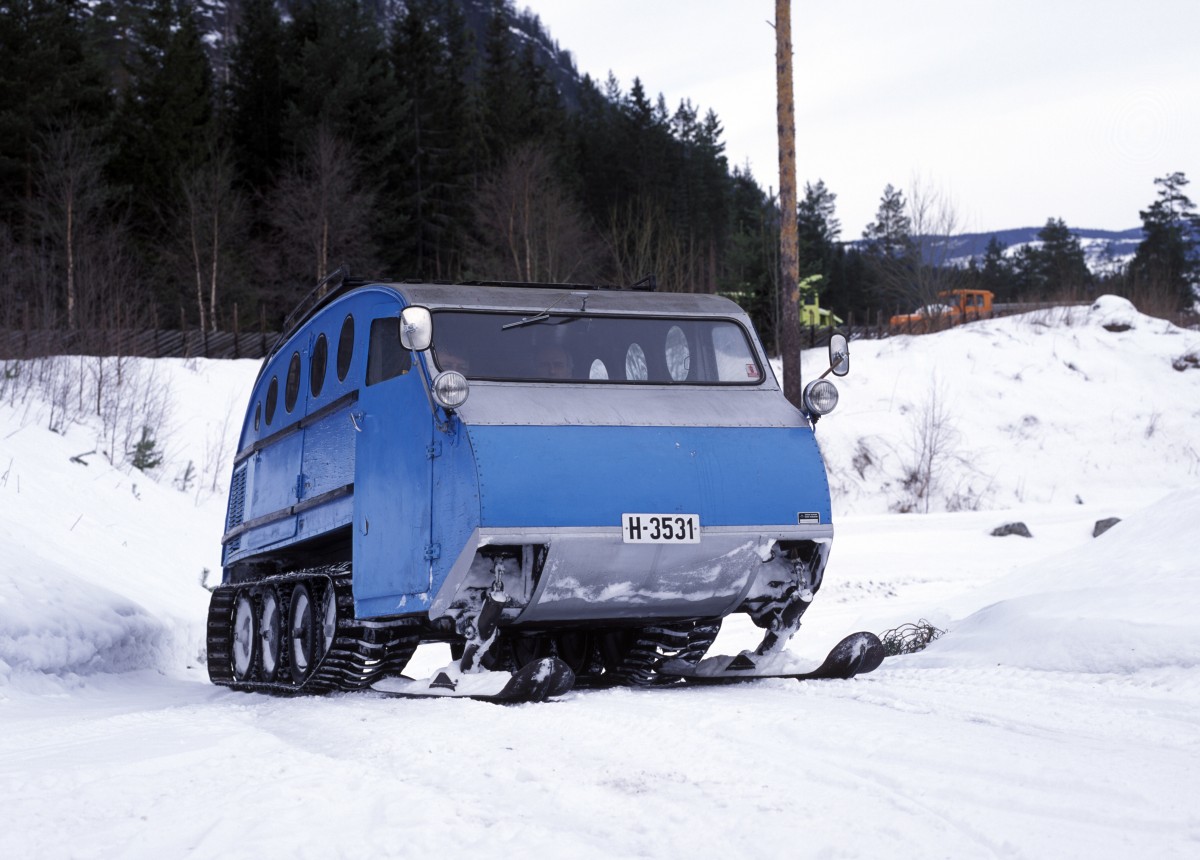 Blå beltebil på snødekt vei, omgitt av trær i vinterlandskap. Snøscooterkjøring i skogen.