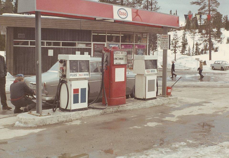 Esso bensinstasjon om vinteren, med bil og snø rundt, noen fyller bensin. Retro stil.