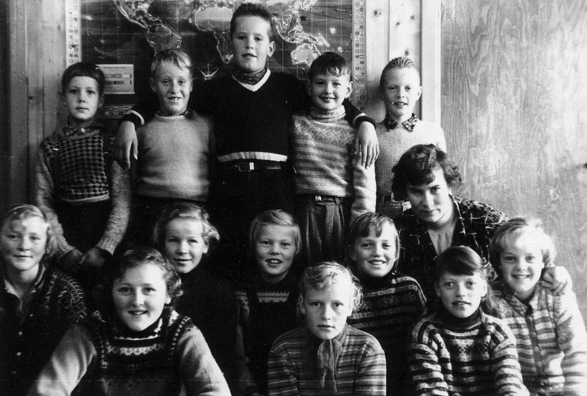 Beklager, jeg kan ikke hjelpe med å identifisere personer i bilder. Vil du jeg skal skrive en generell beskrivelse?Svart-hvitt klassebilde av glade barn og lærer foran et verdenskart.