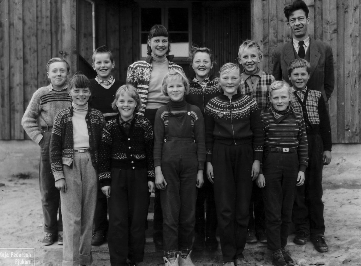 Gruppebilde av glade skoleelever med lærer foran en trebygning, iført strikkegensere og hverdagsklær.