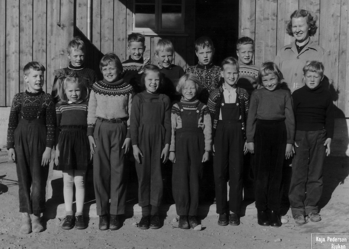 Svart-hvitt bilde av en skoleklasse med barn i tradisjonelle gensere og lærer foran en trebygning.