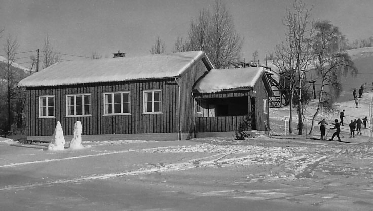 Gammelt trehus om vinteren nær skiløype, omgitt av snø. Skiløpere i bakgrunnen nyter solfylt vinterlandskap.