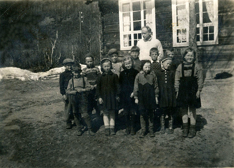 Gammelt klassebilde fra en norsk skole med lærer og elever foran en trebygning, vårstemning i bakgrunnen.