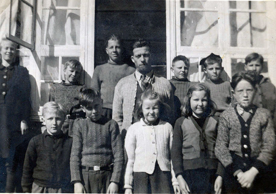 Gammeldags gruppebilde av barn og en mann foran et hus, alle i varme klær, smiler svakt mot kameraet.
