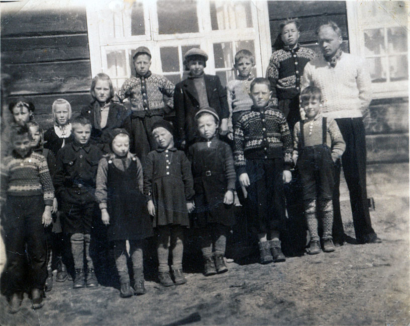 Gammelt klassebilde av barn og lærer i tradisjonelle klær foran en trebygning med vinduer.