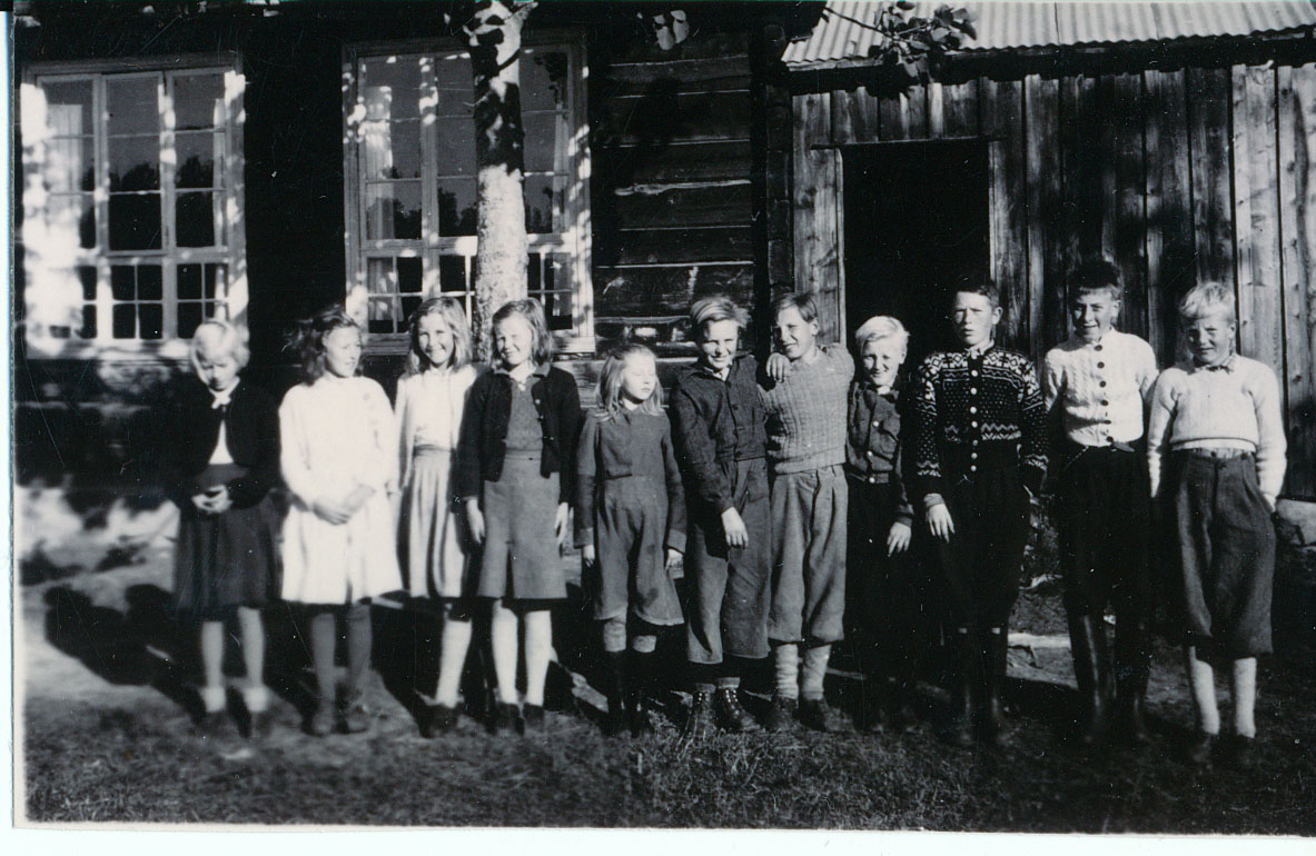 Svart-hvitt bilde av en gruppe barn i klær fra midten av 1900-tallet foran en tømmerbygning.