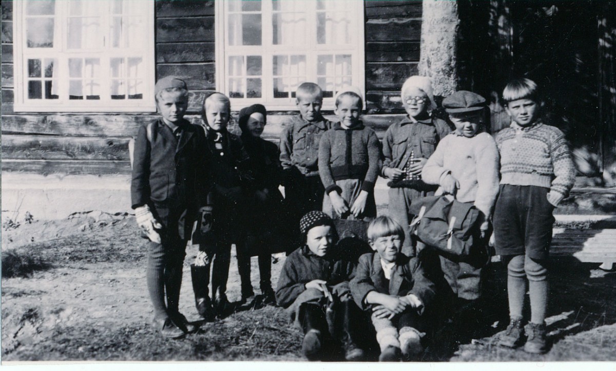 Gamle svart-hvitt bilde av barn utenfor en trebygning, iført typiske 1900-tallsklær.