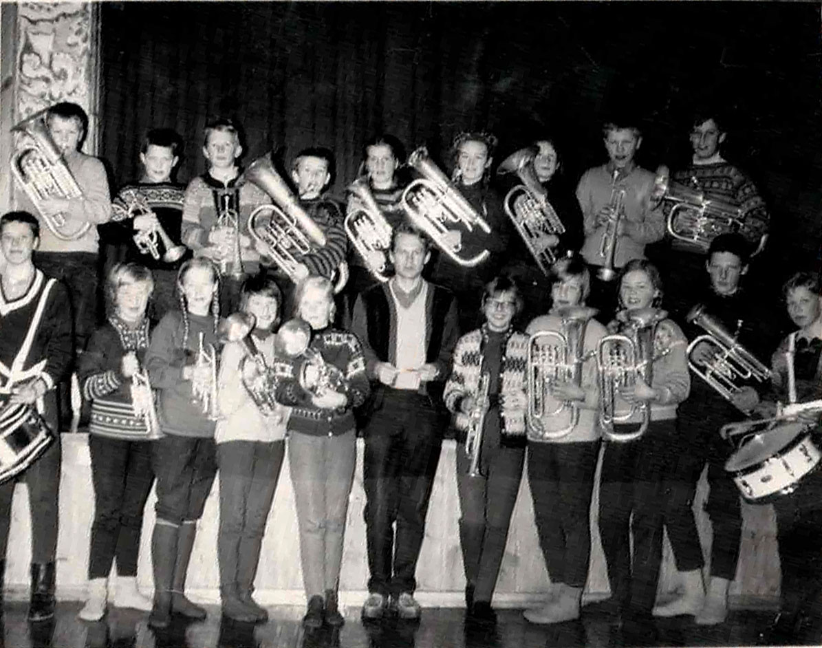 En gruppe unge musikere i uniform spiller ulike blåseinstrumenter på en scene. Vintage svart-hvitt foto.
