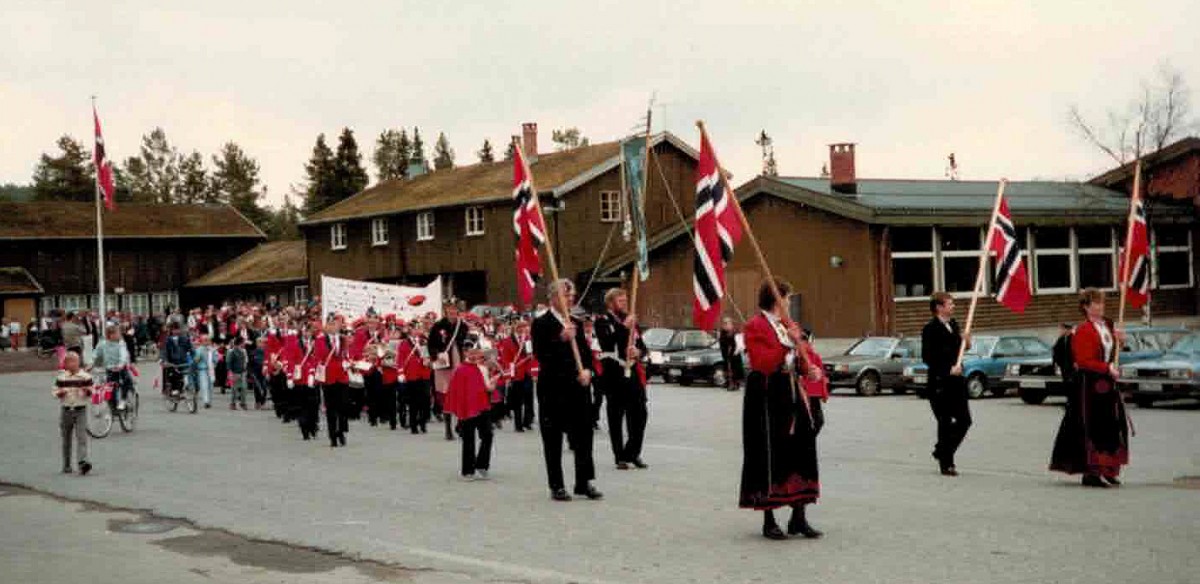 Nasjonal parade med norske flagg og bunader i en bygdegate, 17. mai-feiring med korps og syklister.