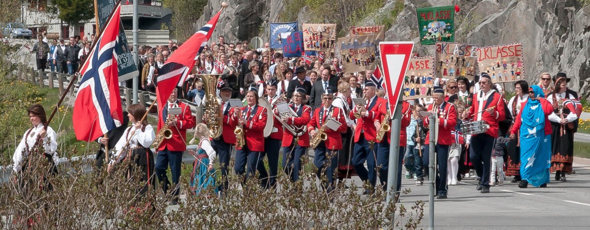 Norsk nasjonaldag parade med musikere i røde uniformer og flagg. Folkemengde i bakgrunnen, mai-feiring.
