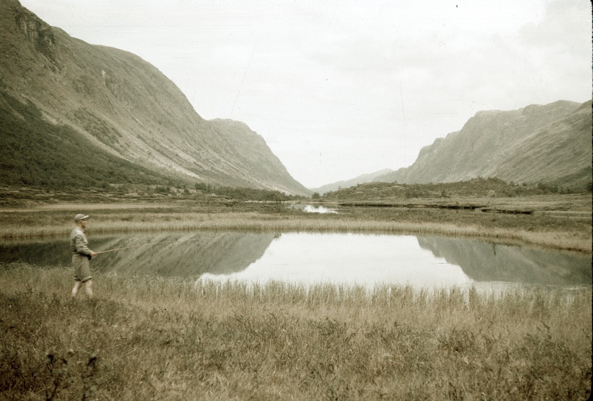 Fisker ved speilblank innsjø i norsk fjellandskap, omgitt av majestetiske fjell og rolig natur.