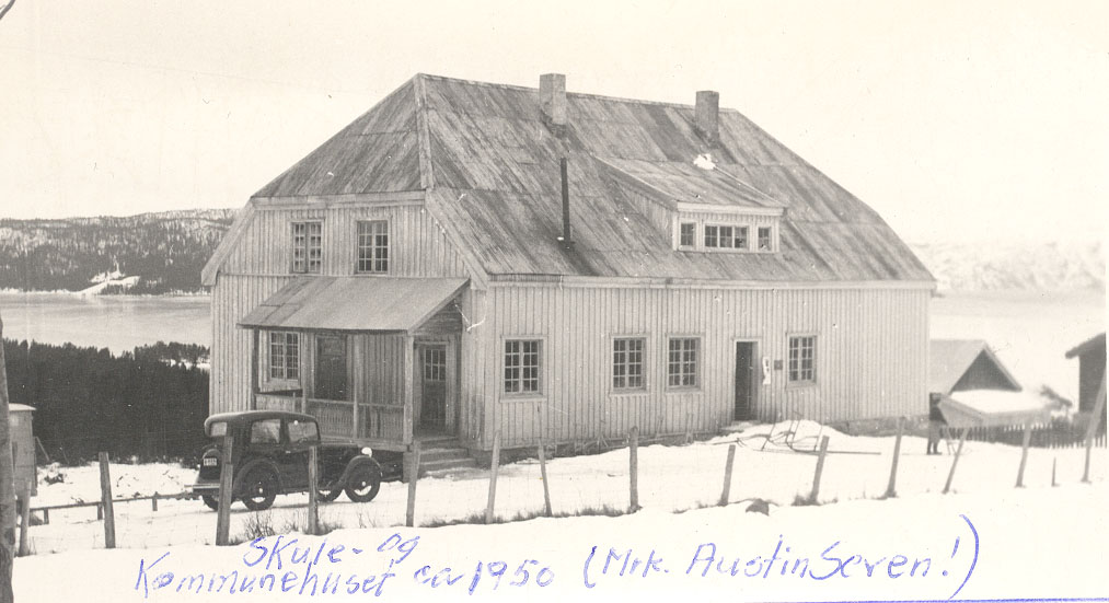 Gammelt skole- og kommunehus med bil i snødekt landskap ved fjord. Historisk trebygning, år circa 1950.
