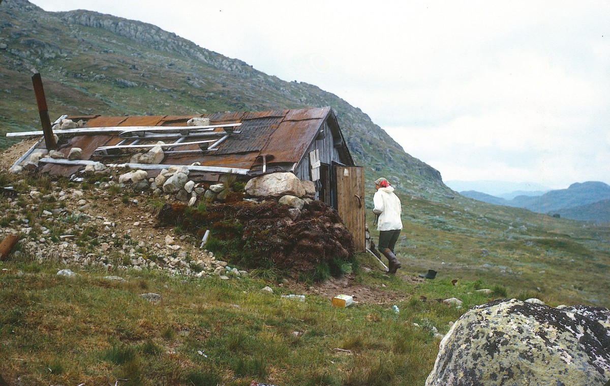 Mann ved en gammel fjellhytte i et norsk landskap, med steiner på taket og fjell i bakgrunnen.